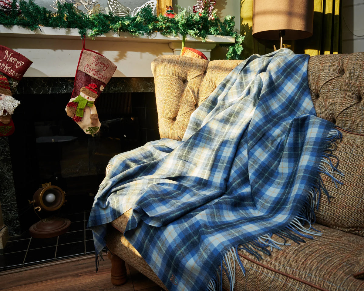 A blue and white plaid fringed throw blanket is draped over a tufted beige armchair in a cozy living room setting with a fireplace, Christmas stocking, and holiday garland.