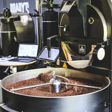 A large black coffee roasting machine with a rotating drum filled with roasted coffee beans, shown in a workshop or production setting with a laptop and control screens nearby.