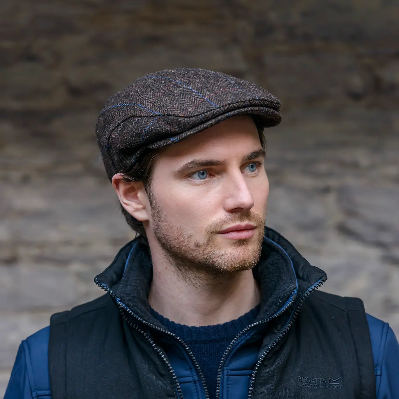 Man wearing a tweed brown flat cap and dark jacket against a stone wall.