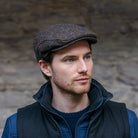 Man wearing a tweed brown flat cap and dark jacket against a stone wall.