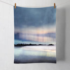 A rectangular kitchen towel or tea towel printed with an abstract seascape scene, hanging from two wooden clothespins on a white string against a white wall.