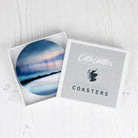 Round coaster shown on a white wood surface, one open to reveal a blue coastal landscape coaster and one closed with a gray lid printed with the text 'Cath Waters Scottish Landscape Coasters' and a map graphic.