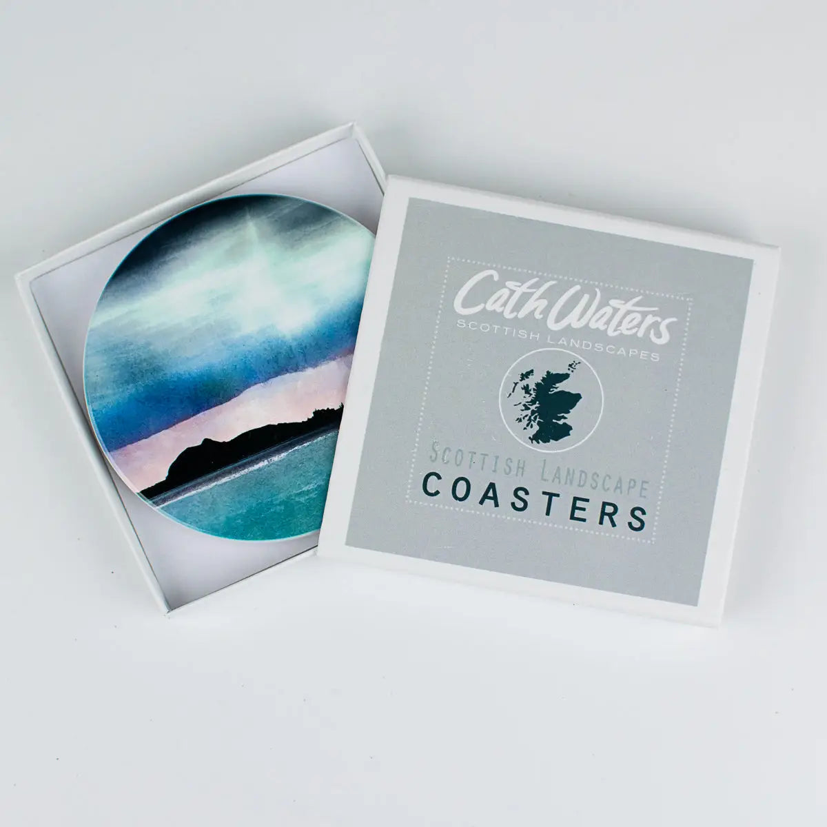 A square product box labeled 'Cath Waters Scottish Landscape Coasters' with one coaster partially slid out, showing a circular printed coaster design of a coastal landscape in blue, teal, black, and pale pink tones.