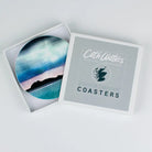 A square product box labeled 'Cath Waters Scottish Landscape Coasters' with one coaster partially slid out, showing a circular printed coaster design of a coastal landscape in blue, teal, black, and pale pink tones.