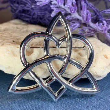 A polished pewter metal Celtic knot triquetra brooch with a pin back, displayed on a blue textured surface with a beige stone and purple flowers in the background.