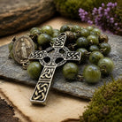 A green stone bead rosary with an ornate silver Celtic cross and a medallion, arranged on a stone slab with moss and dried flowers.