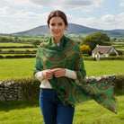 A woman standing outdoors in a green rural landscape, wearing a green shawl with a gold and dark paisley pattern over a white sweater and blue jeans.