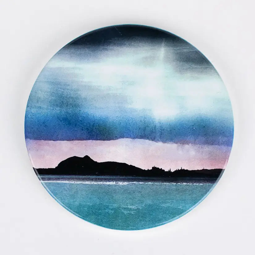 A ceramic cork coaster featuring a watercolor landscape print of a dark mountain silhouette against a blue and teal sky with a soft pink horizon, shown on a plain white background.