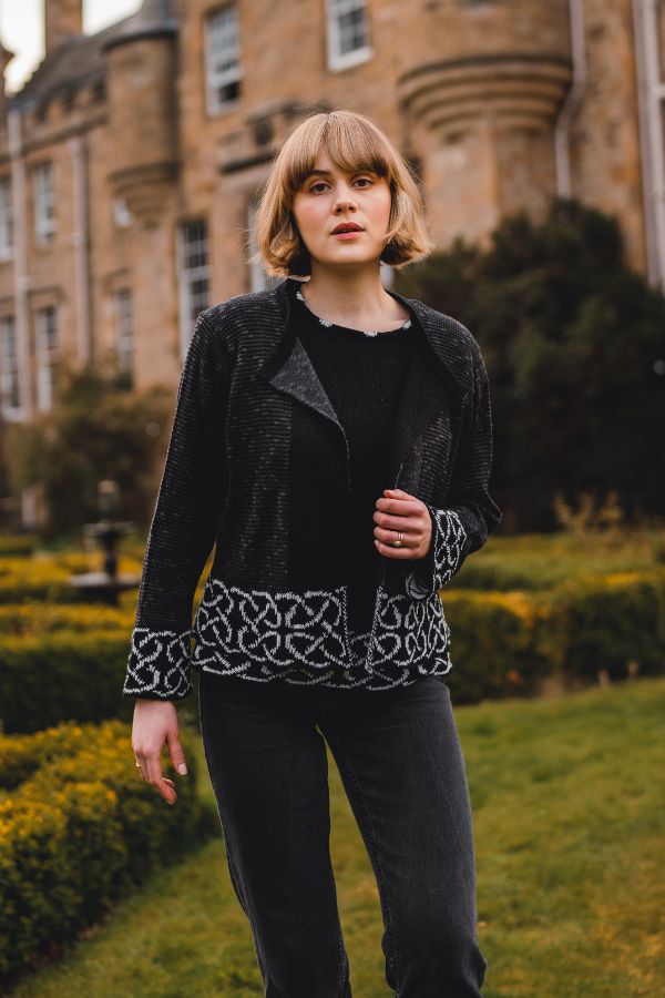 Woman wearing a black jacket with intricate patterns in an outdoor setting with a castle-like building.