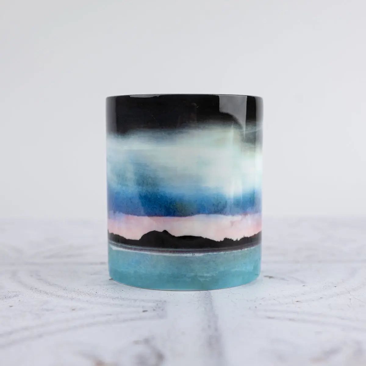 A ceramic mug painted scenic landscape design, featuring a black sky, blue and white clouds, a pink horizon, and a turquoise sea with black mountain silhouettes, set against a light gray background.