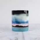 A ceramic mug painted scenic landscape design, featuring a black sky, blue and white clouds, a pink horizon, and a turquoise sea with black mountain silhouettes, set against a light gray background.