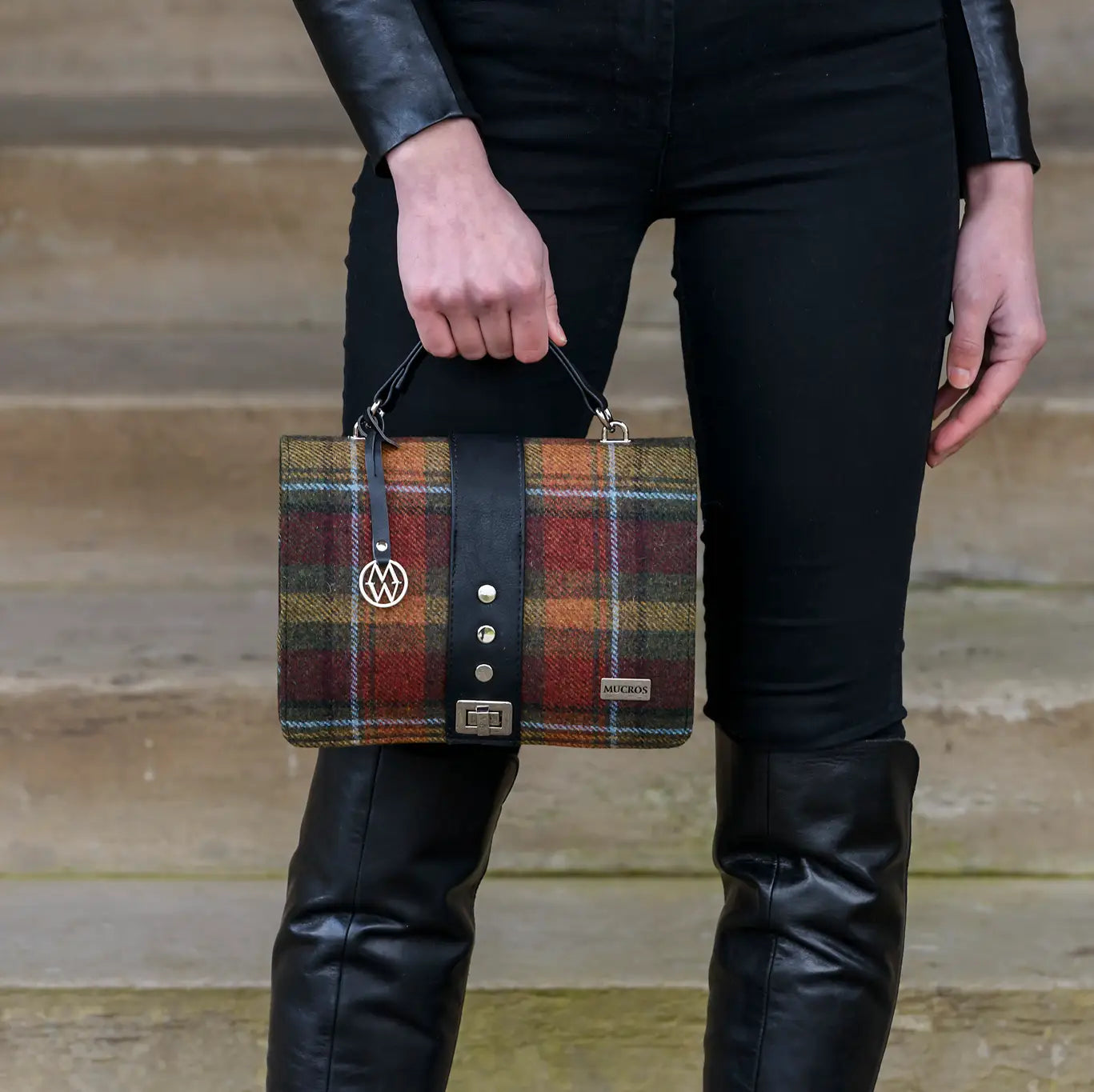 A person wearing black leather boots and a black leather jacket holds a plaid tweed briefcase-style handbag with a black leather strap and silver hardware, standing against a wooden step background.