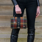 A person wearing black leather boots and a black leather jacket holds a plaid tweed briefcase-style handbag with a black leather strap and silver hardware, standing against a wooden step background.