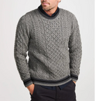 A man wearing a gray cable-knit crewneck sweater with dark ribbed trim, paired with dark trousers, against a light gray studio background.