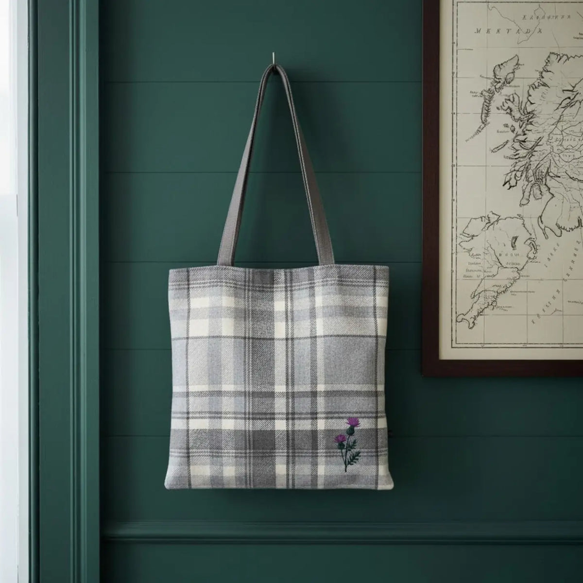 A gray and white plaid fabric tote bag with brown shoulder straps hangs on a dark green paneled wall. A small purple flower embroidery is visible near the bottom right corner of the bag.