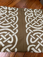 A close-up of a woven textile with a repeating interlocking knot pattern in white over a medium brown background, laid flat on a wooden surface.