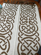 A woven throw blanket with a cream background and a bold brown Celtic knot pattern, laid flat on a wooden table with fringe visible at the top.