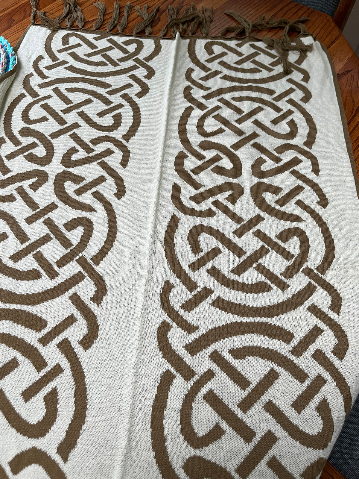 A woven throw blanket with a cream background and a bold brown Celtic knot pattern, laid flat on a wooden table with fringe visible at the top.