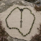 A beaded rosary with olive green stone beads and a silver-toned cross pendant, arranged on a beige linen cloth over a natural rock surface.