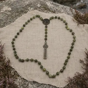A beaded rosary with olive green stone beads and a silver-toned cross pendant, arranged on a beige linen cloth over a natural rock surface.
