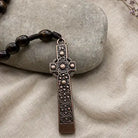 A close-up of a dark brown beaded necklace with an ornate, engraved bronze-toned cross pendant, resting on a textured beige fabric surface next to a smooth gray stone.