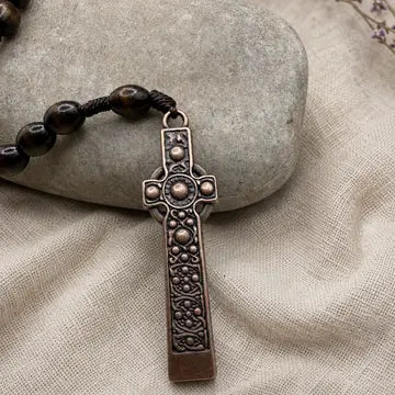 A close-up of a dark brown beaded necklace with an ornate, engraved bronze-toned cross pendant, resting on a textured beige fabric surface next to a smooth gray stone.