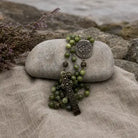 A green beaded rosary with an antique bronze cross and medallion, draped over a smooth gray stone on a beige linen cloth with rocks in the background.