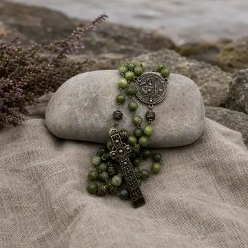 A green beaded rosary with an antique bronze cross and medallion, draped over a smooth gray stone on a beige linen cloth with rocks in the background.