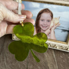 Green shamrock ornament held by a hand with a framed photo of a girl holding a shell in the background.