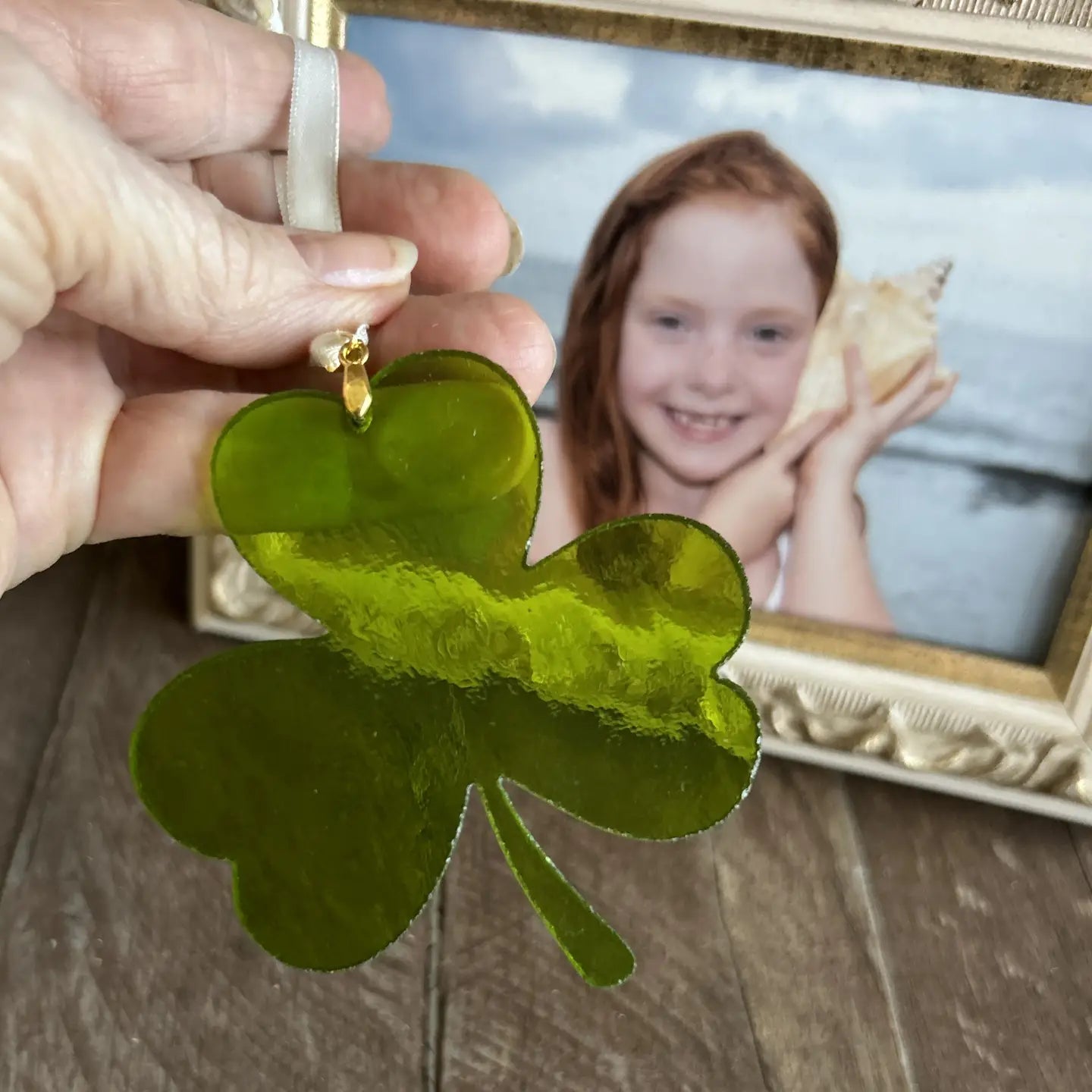 Green shamrock ornament held by a hand with a framed photo of a girl holding a shell in the background.