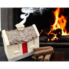 A miniature house-shaped fireplace crackle or decorative stove piece with a textured brown roof, red door, and white base, shown with several brown crackle pieces in front and smoke rising from the top, set against a blurred fireplace with flames.