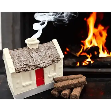 A miniature house-shaped fireplace crackle or decorative stove piece with a textured brown roof, red door, and white base, shown with several brown crackle pieces in front and smoke rising from the top, set against a blurred fireplace with flames.