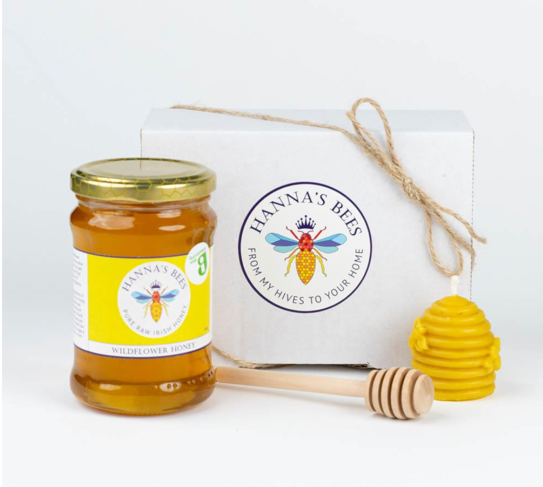 Jar of honey with Hanna's Bees label, honey dipper, and yellow beehive-shaped container on a white background.
