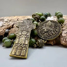 A religious pendant set featuring an antique bronze Orthodox cross with saint engravings, a matching round medallion, and green stone beads, arranged on a piece of bark.
