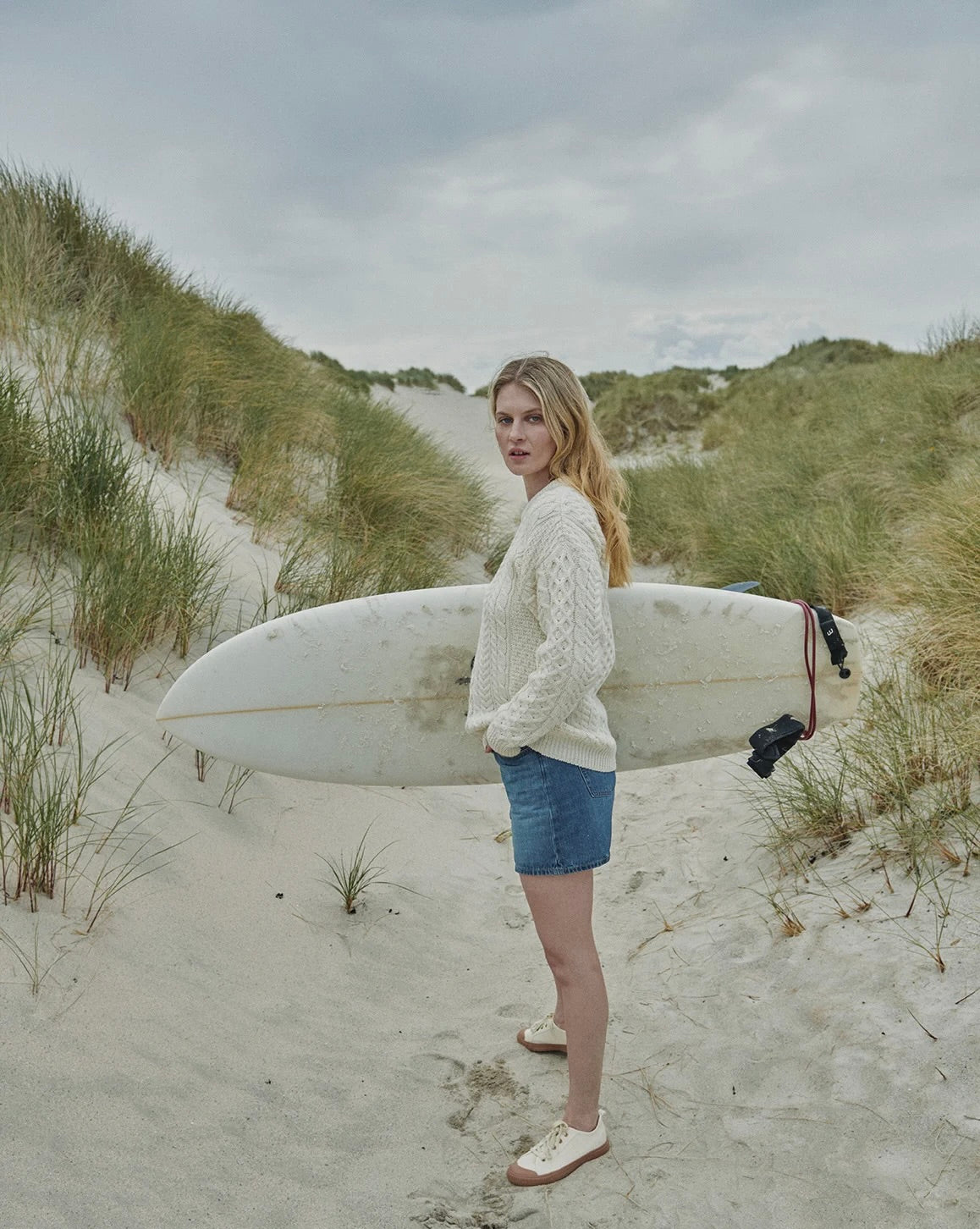 A woman standing on a sandy beach dune holding a white surfboard under her arm, wearing a cream cable-knit sweater, denim shorts, and white sneakers, with beach grass and cloudy skies in the background.
