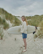 A woman standing on a sandy beach dune holding a white surfboard under her arm, wearing a cream cable-knit sweater, denim shorts, and white sneakers, with beach grass and cloudy skies in the background.