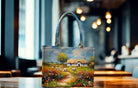 Handbag with a scenic painting of a cottage and field on a wooden table.