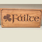 A rectangular wooden plaque with the Irish word 'Fáilte' carved into the center, alongside a carved shamrock. The plaque is made of light brown wood with darker brown engraved details, resting on a light beige surface against a neutral gray wall.