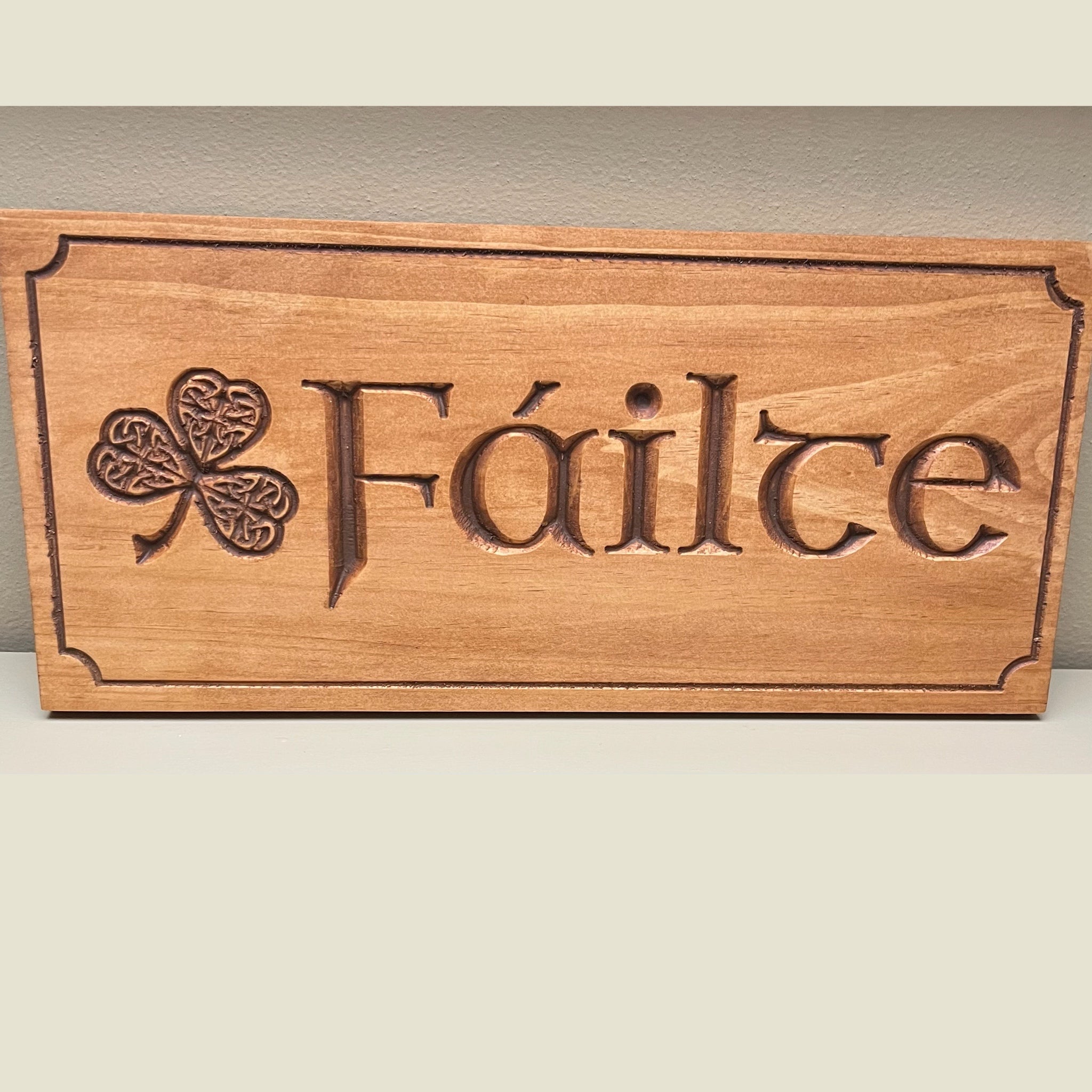 A rectangular wooden plaque with the Irish word 'Fáilte' carved into the center, alongside a carved shamrock. The plaque is made of light brown wood with darker brown engraved details, resting on a light beige surface against a neutral gray wall.