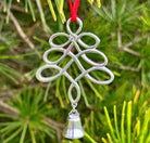 Decorative silver tree-shaped ornament with a bell on a red string, hanging against a green foliage background.