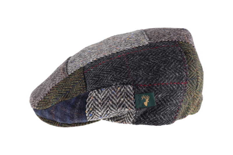 A patchwork herringbone flat cap in a mix of gray, brown, and blue tweed fabrics, featuring a small green embroidered deer logo patch on the side.