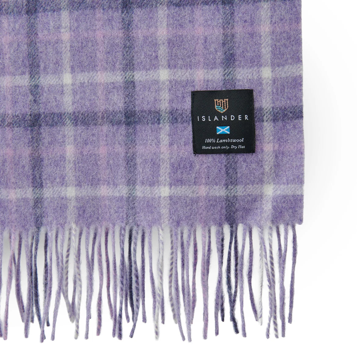 A close-up of a purple and white plaid fringed scarf with a black 'ISLANDER' brand label sewn onto one corner.