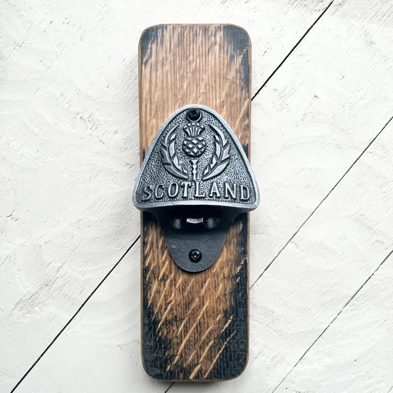 A rustic wall-mounted bottle opener featuring a distressed wood block with a weathered metal Scotland thistle emblem, shown against a white painted wood plank background.