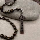A close-up of a rosary necklace with dark brown wooden beads, an ornate dark metal crucifix pendant, and a round medallion, arranged on a light gray stone over a beige fabric background.