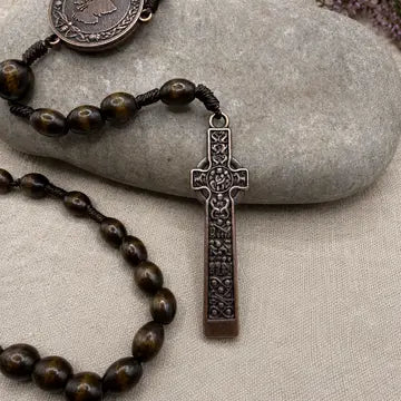 A close-up of a rosary necklace with dark brown wooden beads, an ornate dark metal crucifix pendant, and a round medallion, arranged on a light gray stone over a beige fabric background.