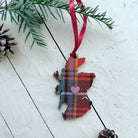 A Scotland map-shaped fabric ornament in a red tartan plaid pattern with a small pink heart in the center, hanging from a red ribbon on a white wooden surface with pine branches and pinecones.
