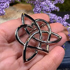 A polished pewter Celtic triquetra knot brooch held in a person's hand, with blurred purple flowers in the background.