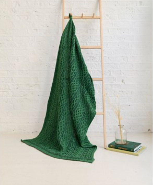 Green knitted blanket draped over a wooden ladder against a white brick wall.