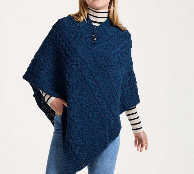 A woman wearing a navy blue cable-knit poncho with a buttoned collar over a black and white striped turtleneck and blue jeans, photographed against a plain light background.