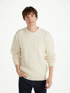 A man wearing a cream-colored cable-knit crewneck sweater and dark blue jeans, standing against a plain white studio background.